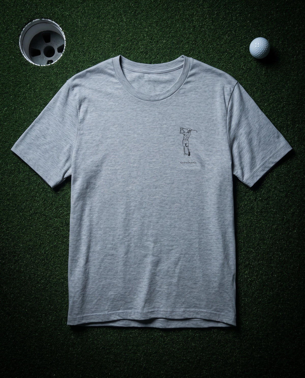The Bones Tee in Ash — front, Bones chest mark, laid flat on a dark putting green at night