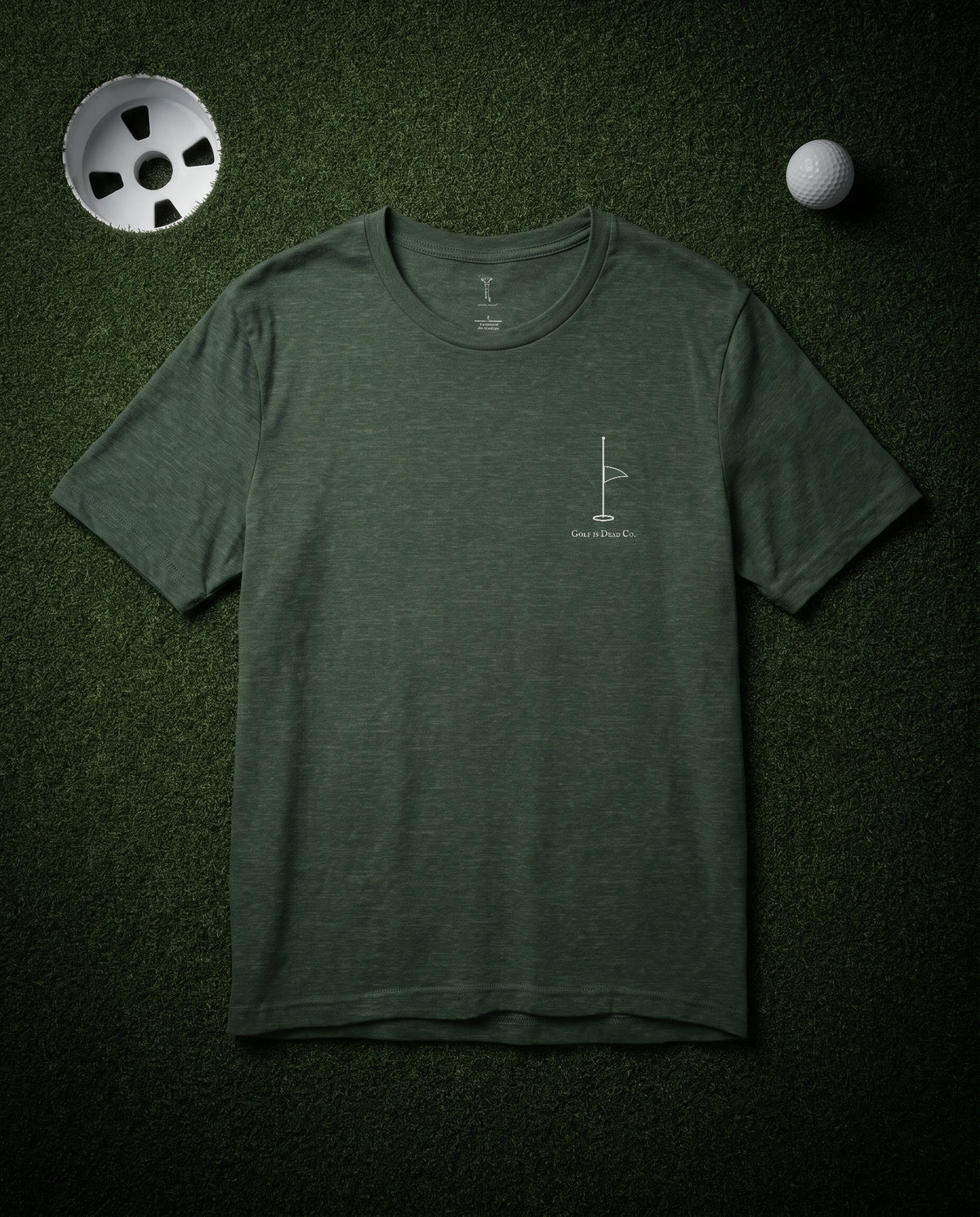 The Last Flag Tee in Fairway — front, half-mast pin flag chest mark, laid flat on a dark putting green at night