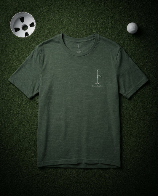 The Last Flag Tee in Fairway — front, half-mast pin flag chest mark, laid flat on a dark putting green at night